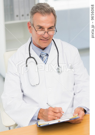 Smiling doctor sitting at his desk writing on clipboard Smiling doctor sitting at his desk writing on clipboard 9656068