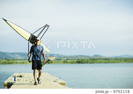 A middle-aged man carrying oars and a rowing shell on his shoulders, on a pontoon. A middle-aged man carrying oars and a rowing shell on his shoulders, on a pontoon. 9702158