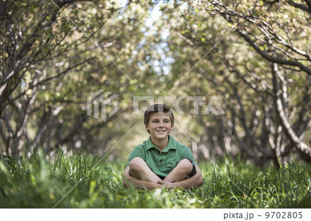 A boy sitting crosslegged in a woodland tunnel with tree branches meeting overhead. 9702805
