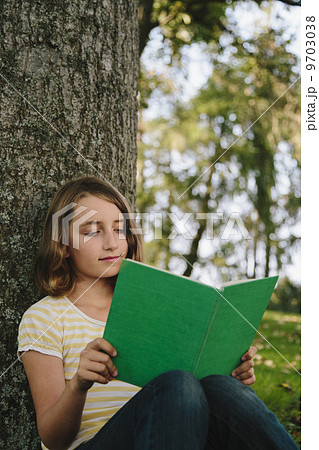 Nine year old girl sitting beneath tree, reading book 9703038