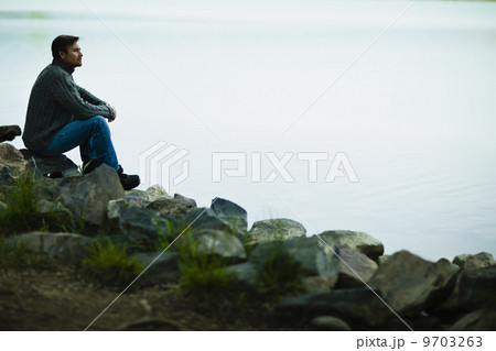 A middle-aged man sitting on rocks looking pensively over the water. 9703263