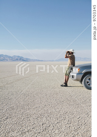 A man leaning against a truck, looking through binoculars. 9703345