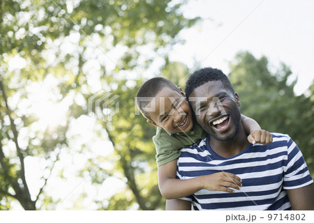A man giving a child a piggyback, in the shade of trees on a summer day. 9714802