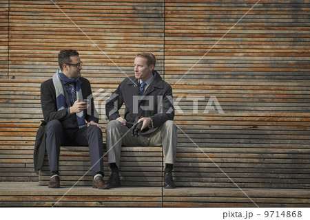 Two men sitting on a bench outside a large building in the city. One checking his mobile phone.  9714868