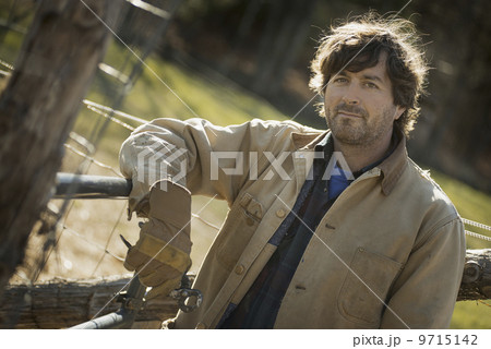 A man in a brown jacket, wearing work gloves leaning on a fence.  Working on an organic farm. 9715142
