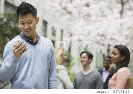 City life in spring. Young people outdoors in a city park. A man checking his cell phone. Four people under a tree in blossom. City life in spring. Young people outdoors in a city park. A man checking his cell phone. Four people under a tree in blossom. 9715315