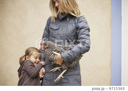A woman in a grey coat holding a black and white chicken with a red coxcomb under one arm. A young girl beside her holding her other hand 9715363