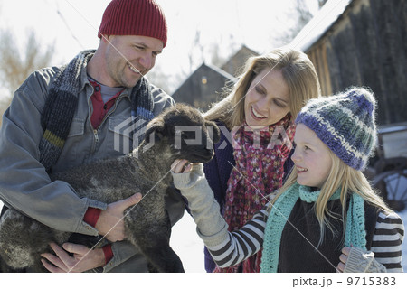 Winter scenery with snow on the ground. A man holding a young lamb, and a child stroking its chin. 9715383