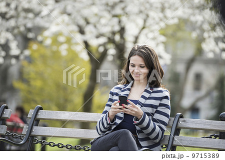 Outdoors in the city in spring time. New York City park. White blossom on the trees. A woman sitting on a bench holding her mobile phone. Outdoors in the city in spring time. New York City park. White blossom on the trees. A woman sitting on a bench holding her mobile phone. 9715389
