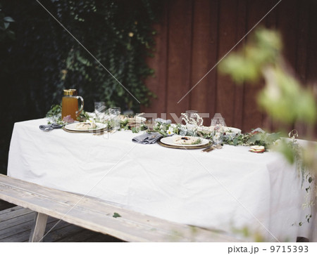 A table laid for a special meal. Place settings, with plates and cutlery. Glasses. A white table cloth and bench seat. Plates of food. 9715393