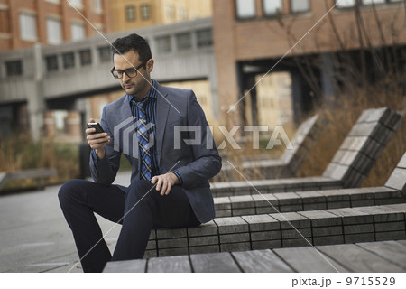 A man in a formal jacket and tie, sitting on a bench outside a city building, checking his phone for messages. A man in a formal jacket and tie, sitting on a bench outside a city building, checking his phone for messages. 9715529