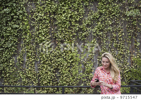 City life in spring. City park with a wall covered in climbing plants and ivy.  A young blonde haired woman checking her smart phone.  9715547