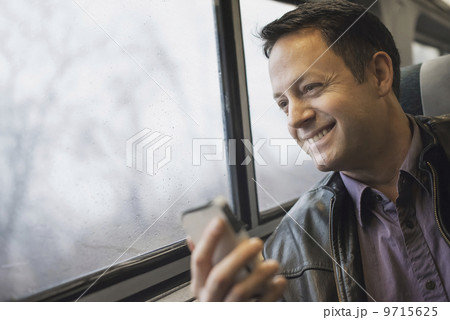 A mature man sitting at a window seat on a train, holding his mobile phone. Smiling and looking in the distance. A mature man sitting at a window seat on a train, holding his mobile phone. Smiling and looking in the distance. 9715625
