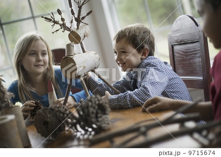 Three children sitting at a table in family home. A twig reindeer in the centre, with branches and twigs to make objects.  9715674