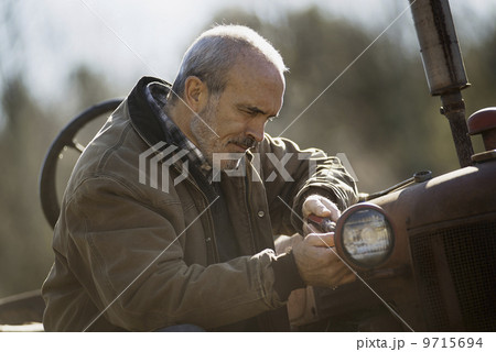 Working on an organic farm. A man in a brown jacket working on farm equipment. A tractor. Working on an organic farm. A man in a brown jacket working on farm equipment. A tractor. 9715694