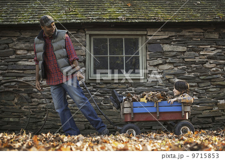 A man in a padded jacket pulling a garden cart with a child sitting among the dried autumn leaves. A traditional stone building. A man in a padded jacket pulling a garden cart with a child sitting among the dried autumn leaves. A traditional stone building. 9715853