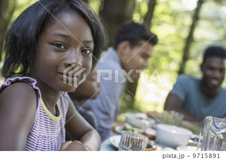 A family picnic meal in the shade of tall trees. A young girl and adults seated at the table. 9715891