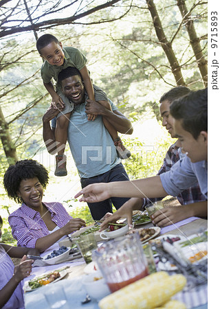 A family picnic meal in the shade of tall trees. A young boy sitting on his father's shoulders. 9715893