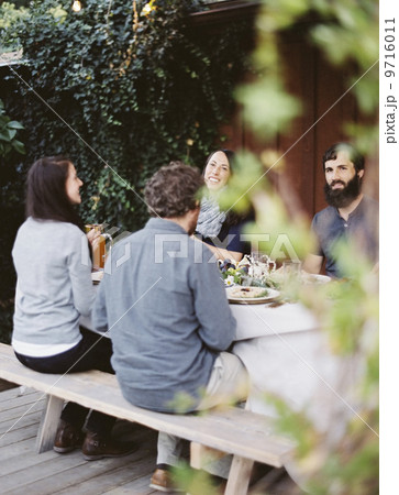 A group of people around a table in a garden. A celebration meal, with table settings and leafy decorations. Glasses plates and cutlery. A group of people around a table in a garden. A celebration meal, with table settings and leafy decorations. Glasses plates and cutlery. 9716011