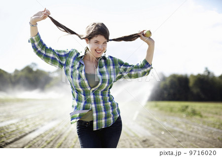 A girl in a green checked shirt with braids standing in a field with sprinklers working in the background.  9716020