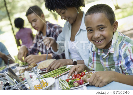 A family picnic meal in the shade of tall trees. Parents and children helping themselves to fresh fruits and vegetables. 9716240