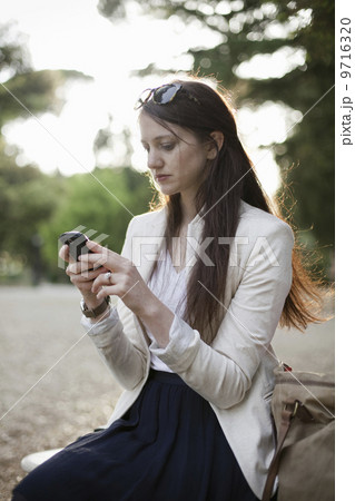 A woman wearing a cream jacket, sitting in a city park looking at her cell phone.  9716320