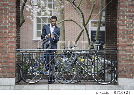 City life in spring. Young people outdoors in a city park. A man in a suit, beside a bicycle park on a sidewalk. Using his smart phone. 9716322