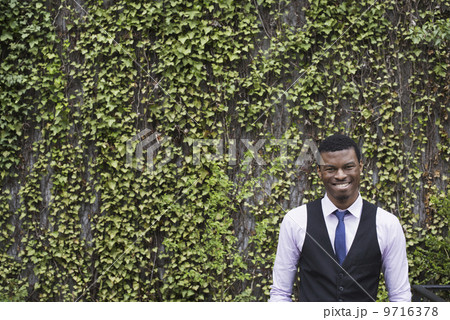 City life in spring. City park with a wall covered in climbing plants and ivy. A young man in a waistcoat, shirt and tie. Looking at the camera. City life in spring. City park with a wall covered in climbing plants and ivy. A young man in a waistcoat, shirt and tie. Looking at the camera. 9716378