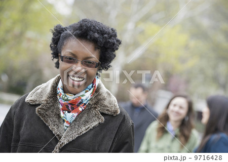 A group of people in a city park. A young woman in a coat with a large collar, smiling and looking at the camera. A group of people in a city park. A young woman in a coat with a large collar, smiling and looking at the camera. 9716428