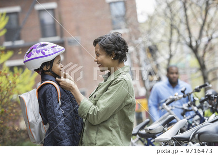 A New York city park in the spring. A boy in a cycle helmet, being fastened by his mother, beside a bicycle rack. 9716473