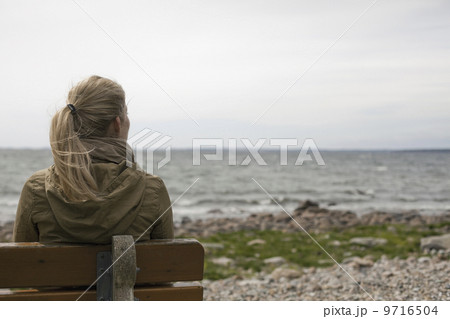 A woman with long blonde hair wearing a brown hooded coat, seated on a bench looking out to sea.  9716504
