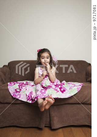 A 3 year old girl with long brown hair in a pink flowered cotton dress with the skirt spread out, sitting on a brown sofa. 9717081