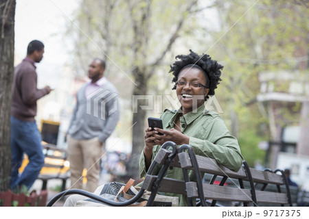 Outdoors in the city in spring. An urban lifestyle. A woman on a bench checking her phone. Two men in the background. Outdoors in the city in spring. An urban lifestyle. A woman on a bench checking her phone. Two men in the background. 9717375