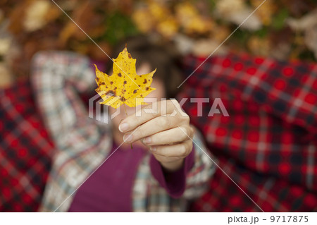 A woman lying on a red tartan picnic blanket, looking upwards, holding a maple leaf. A woman lying on a red tartan picnic blanket, looking upwards, holding a maple leaf. 9717875