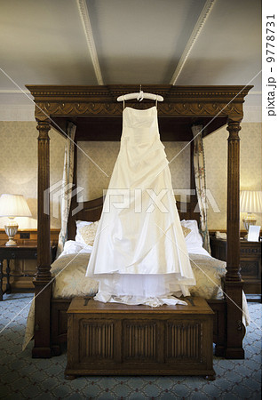 A full length white wedding dress hanging from a hanger on a four-poster bed. A full length white wedding dress hanging from a hanger on a four-poster bed. 9778731