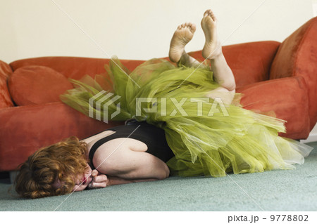 A woman lying on her front on the floor, wearing a lime green tutu, kicking her legs in the air. 9778802