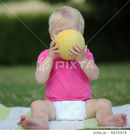 Adorable little girl is sitting on a carpet in a park and biting on a half of melon 9820928