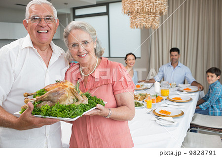 Grandparents holding chicken roast with family at dining table 9828791