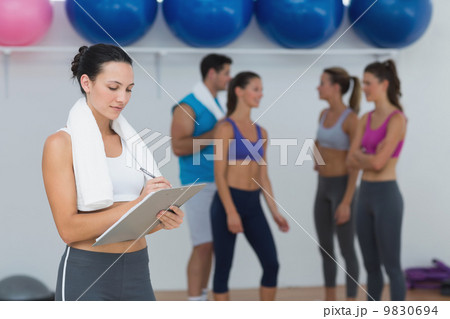 Female trainer writing on clipboard with class in background Female trainer writing on clipboard with class in background 9830694