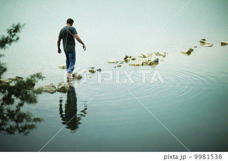 A man walking barefoot across stepping stones away from the shore of a lake. 9981536