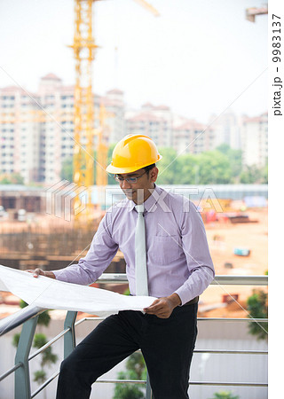 indian male architect reading plans with construction background indian male architect reading plans with construction background 9983137