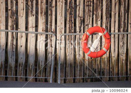 Red lifebuoy with rope on metal railings above weathered wooden wall in port 10096024