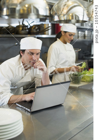 Chef using laptop and cook preparing food in commercial kitchen Chef using laptop and cook preparing food in commercial kitchen 10354743