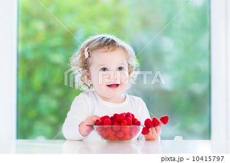 Beautiful little girl eating raspberry in a white dining room at Beautiful little girl eating raspberry in a white dining room at 10415797