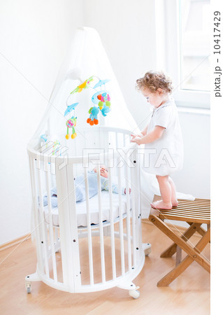 Cute toddler girl standing on a chair and watching her newborn b 10417429