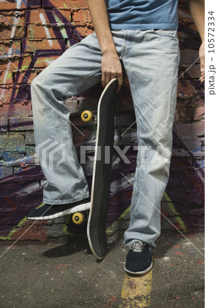 Asian man holding skateboard in front of graffitied wall Asian man holding skateboard in front of graffitied wall 10572334