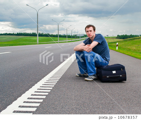 The young man sits pending on road with a suitcase The young man sits pending on road with a suitcase 10678357