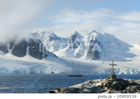 A cross and grave on a hilltop overlooking the snow capped peaks of the mountains on Petterman island. A tall masted ship in the channel.  10909559