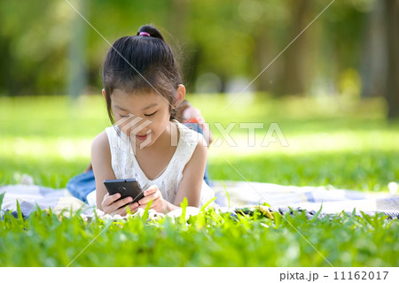 Little girl in causal using mobile smart phone laying on a mat in a park Little girl in causal using mobile smart phone laying on a mat in a park 11162017
