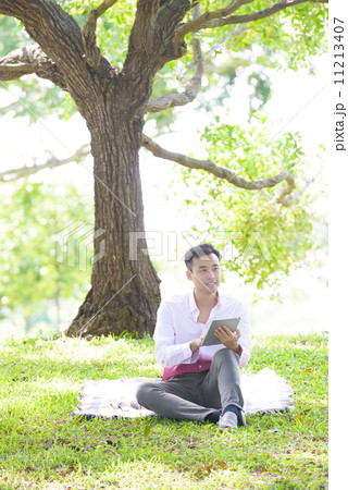 Young man in causal using mobile device tablet iPad sitting on a mat in a park 11213407